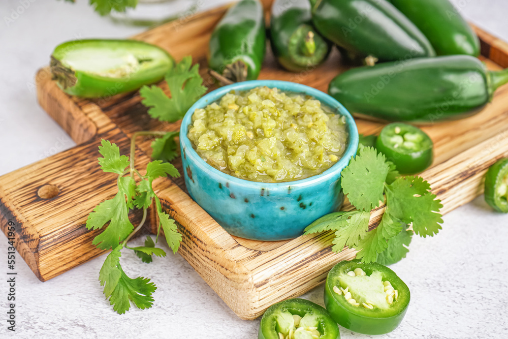 Wooden board with bowl of tasty green salsa sauce, jalapeno peppers and cilantro on light background