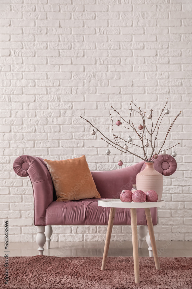 Vase with tree branches, Christmas balls on table and armchair near white brick wall