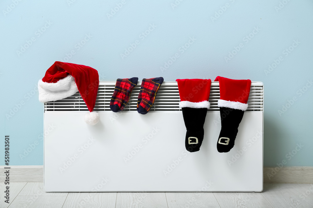 Santa hat, warm socks and baby bodysuit drying on electric radiator near light wall