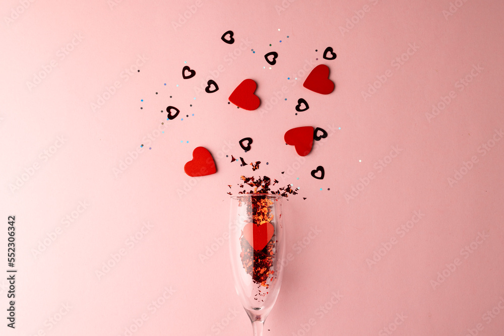 Champagne glass spilling red heart shapes and confetti on pale pink background with copy space