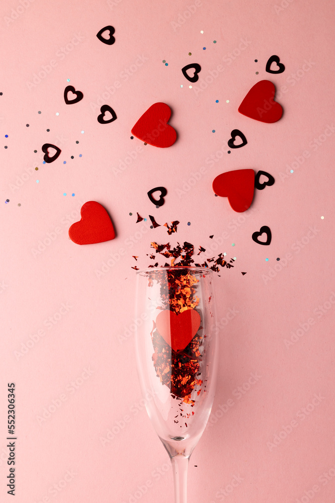 Vertical of champagne glass spilling red heart shapes on pale pink background with copy space