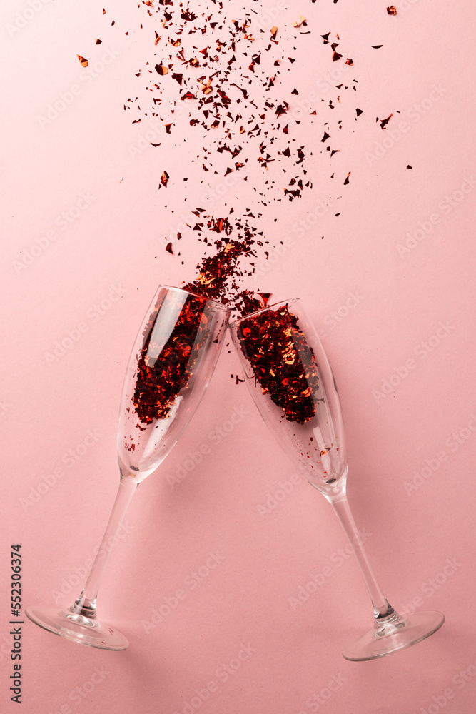 Vertical of two champagne glasses spilling red glitter confetti on pale pink background