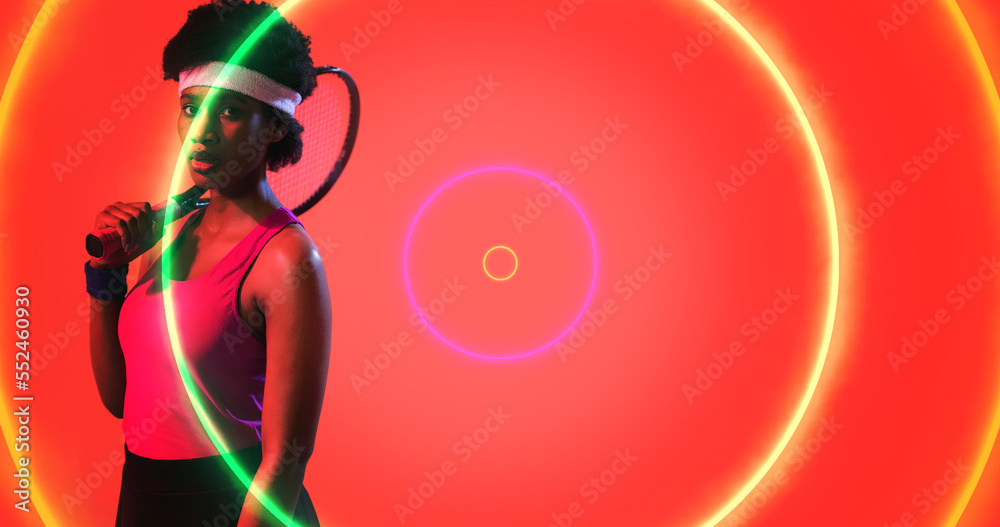 Biracial confident female tennis player holding racket by illuminated circles on red background