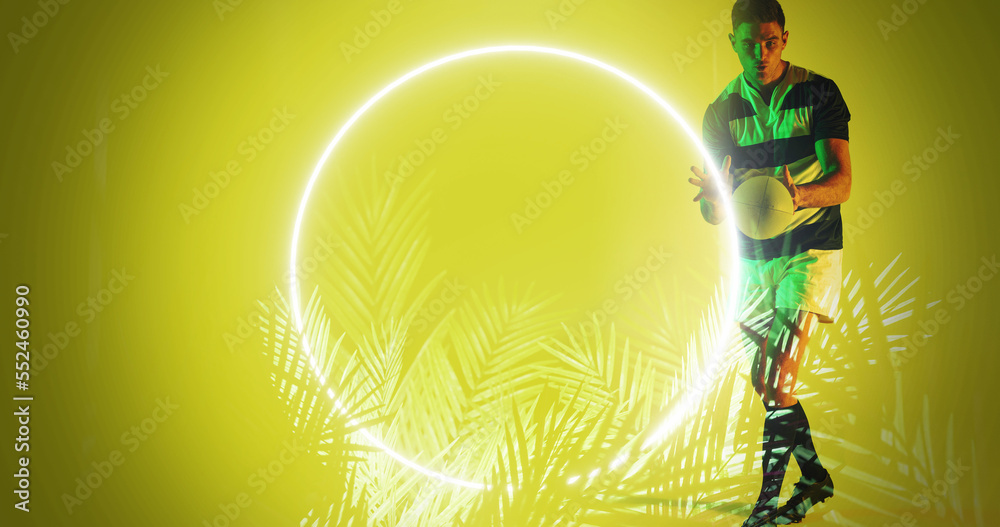 Caucasian male player catching rugby ball by illuminated circle and plants on yellow background