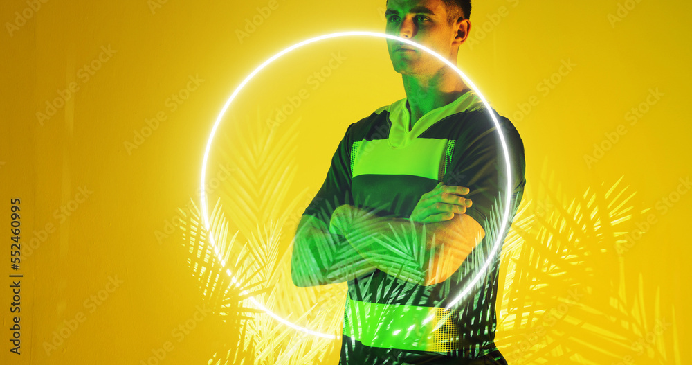 Caucasian rugby player with arms crossed by illuminated circle and plants on yellow background