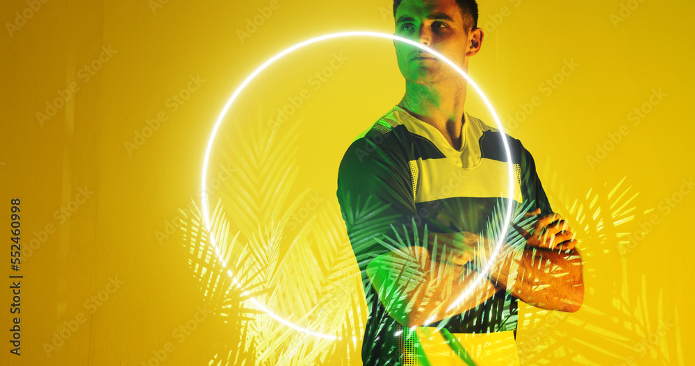 Thoughtful caucasian rugby player with arms crossed by circle and plants on yellow background