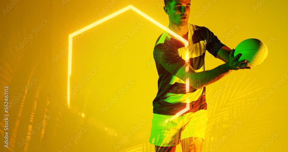 Caucasian male player catching rugby ball by illuminated hexagon and plants on yellow background