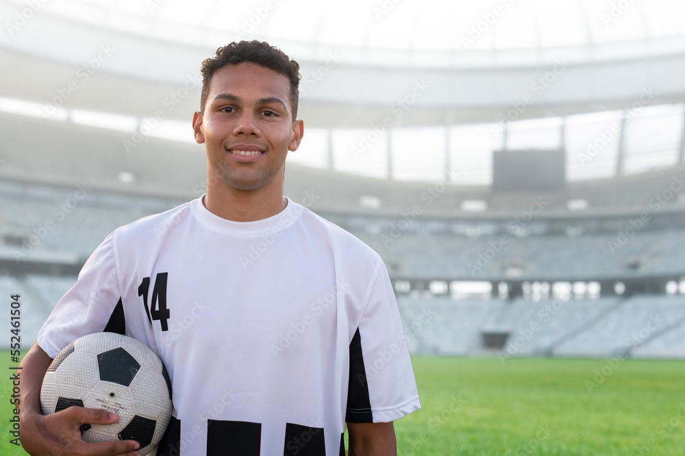 Portrait of smiling biracial young soccer player with ball in stadium on sunny day, copy space