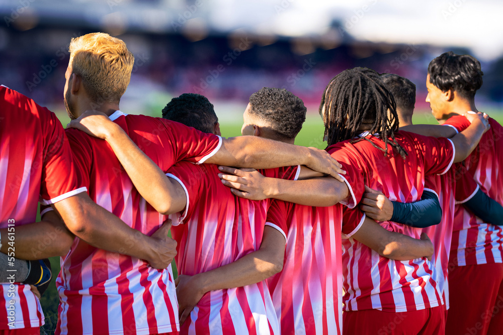 Team of multiracial male players in sports clothing with arms around at soccer field