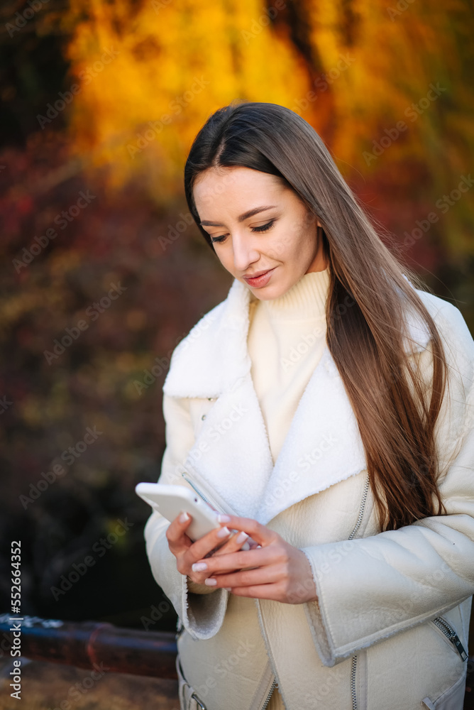 年轻漂亮的女人，秋天公园背景是手机。户外移动的女模特。