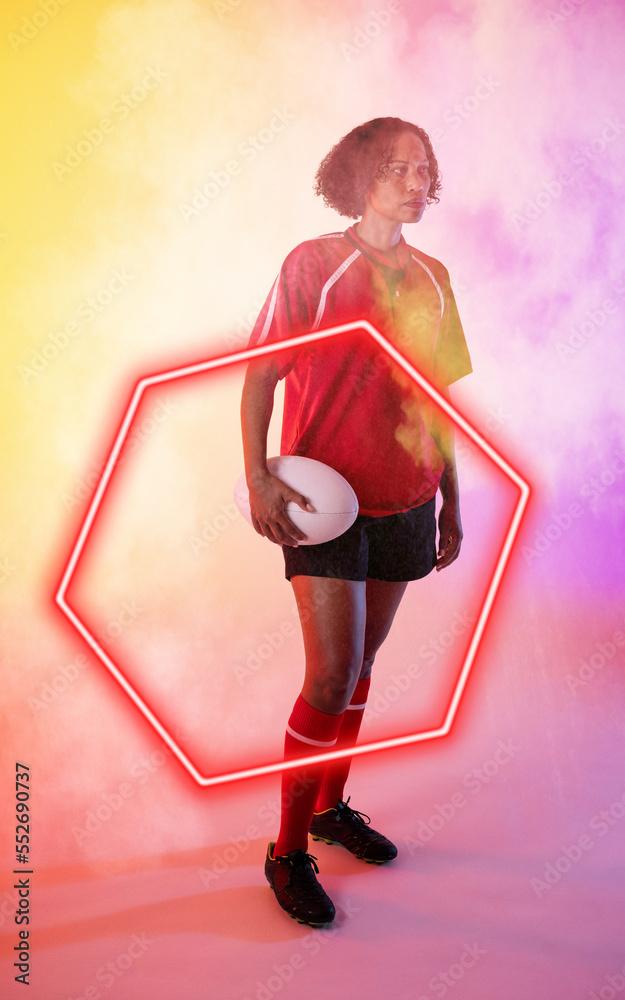 Biracial female rugby player holding ball and standing by illuminated hexagon on smoky background