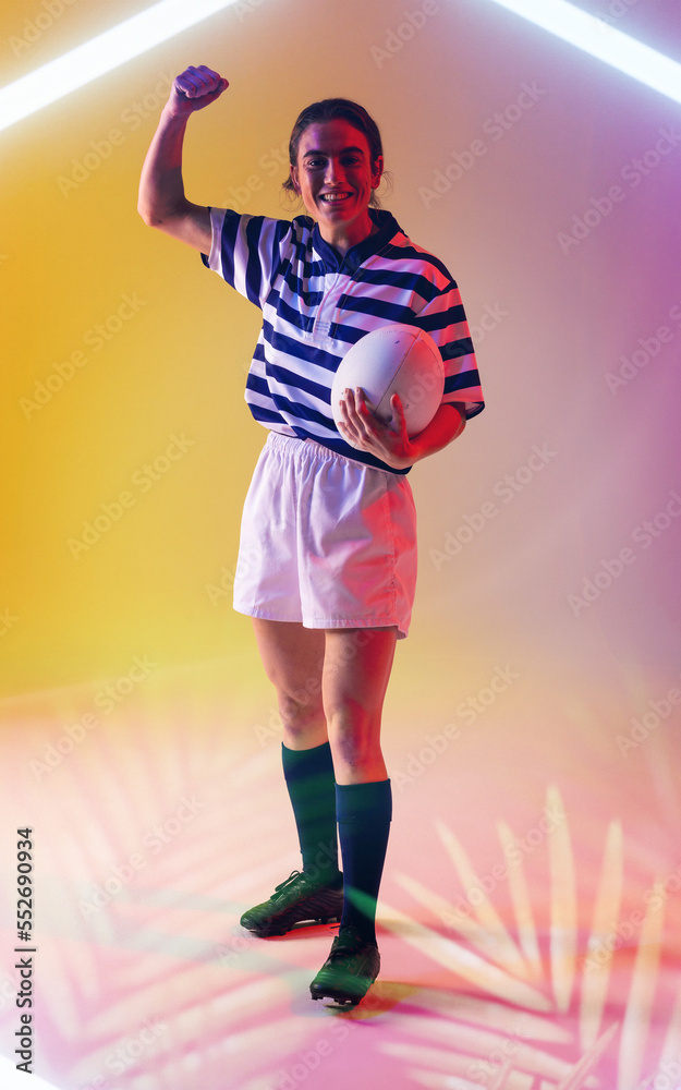 Happy caucasian female rugby player holding ball and raising hand over illuminated lines and plants