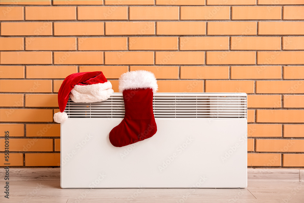 Santa hat and Christmas sock on electric radiator near brick wall