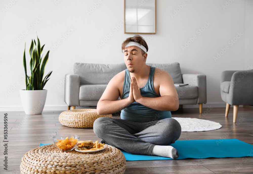 Young overweight man meditating near unhealthy food at home