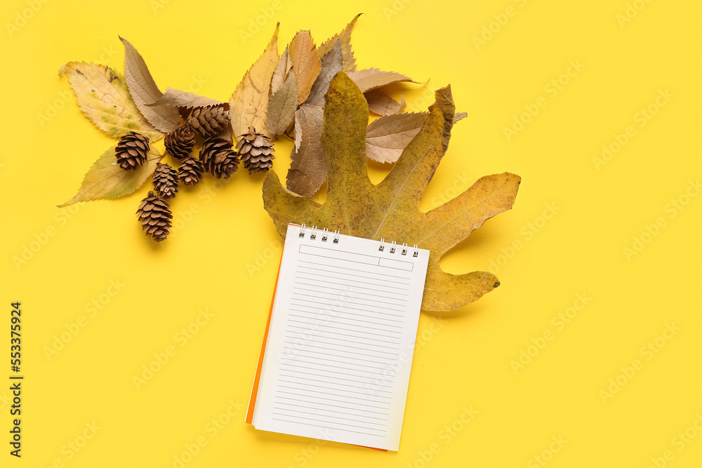 Composition with blank notebook, autumn leaves and cones on yellow background