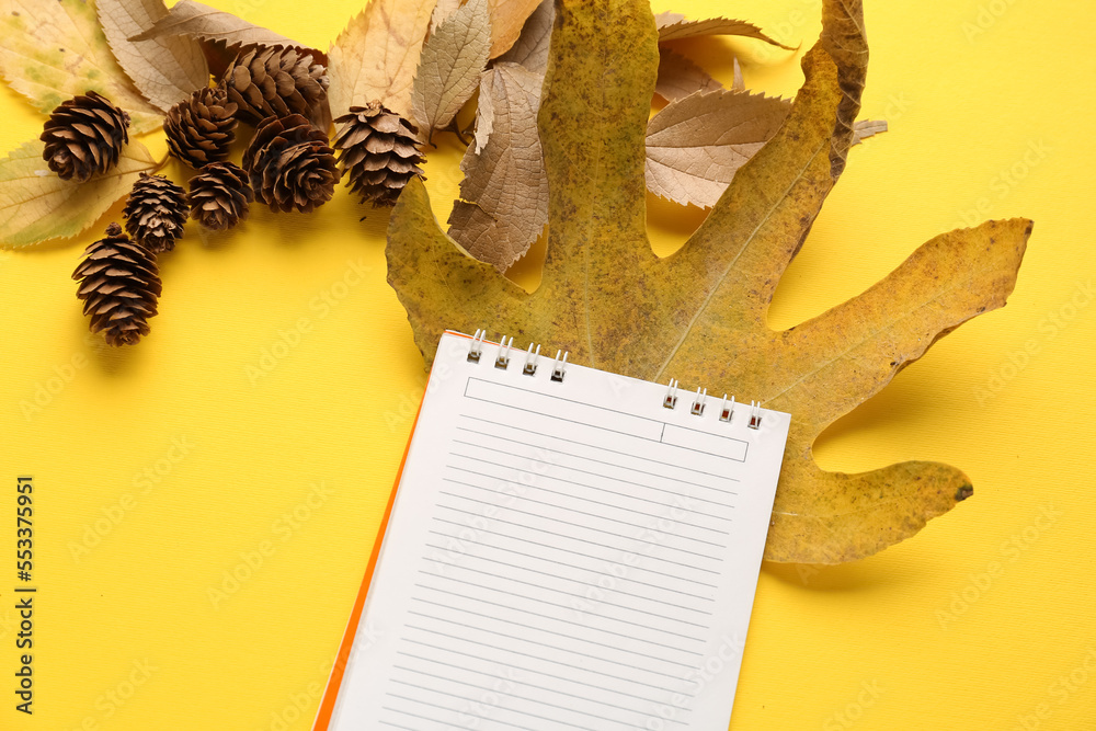 Composition with blank notebook, autumn leaves and cones on yellow background