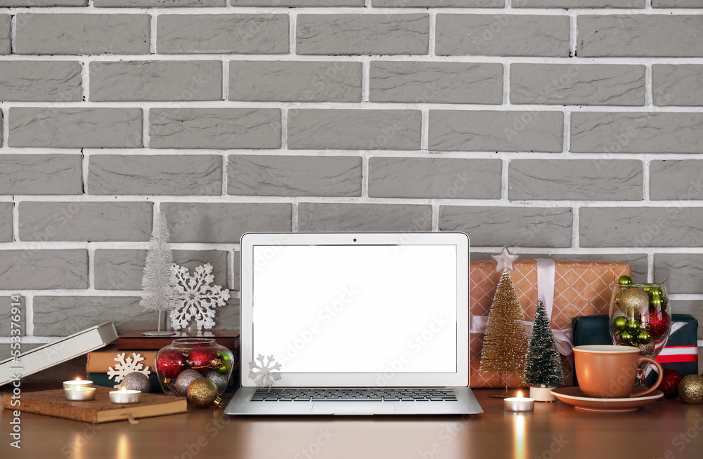 Laptop with cup, Christmas decor and presents on table near grey brick wall