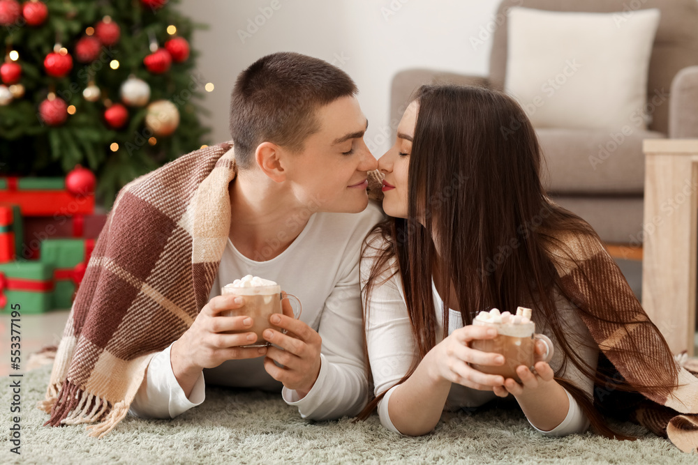 Young couple with plaid and cups of cocoa lying at home on Christmas eve