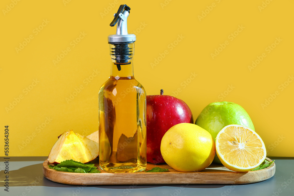 Wooden board with bottle on apple cider vinegar and fruits on table near color wall