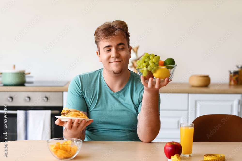 Young overweight man choosing fruits over fast food at table in kitchen