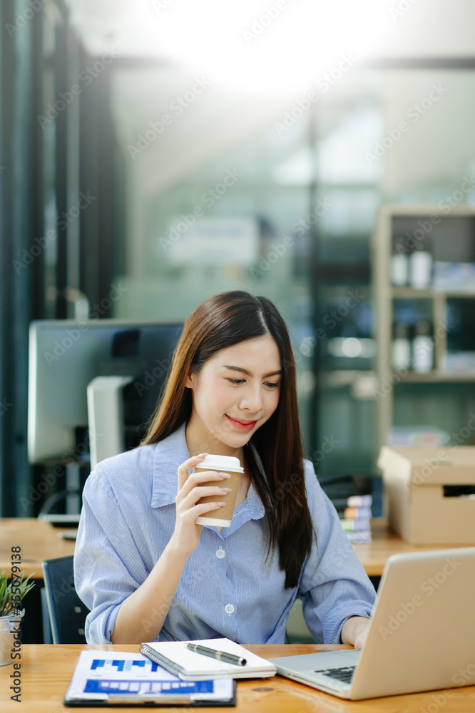 Confident beautiful Asian businesswoman typing laptop computer and digital tablet while holding coff