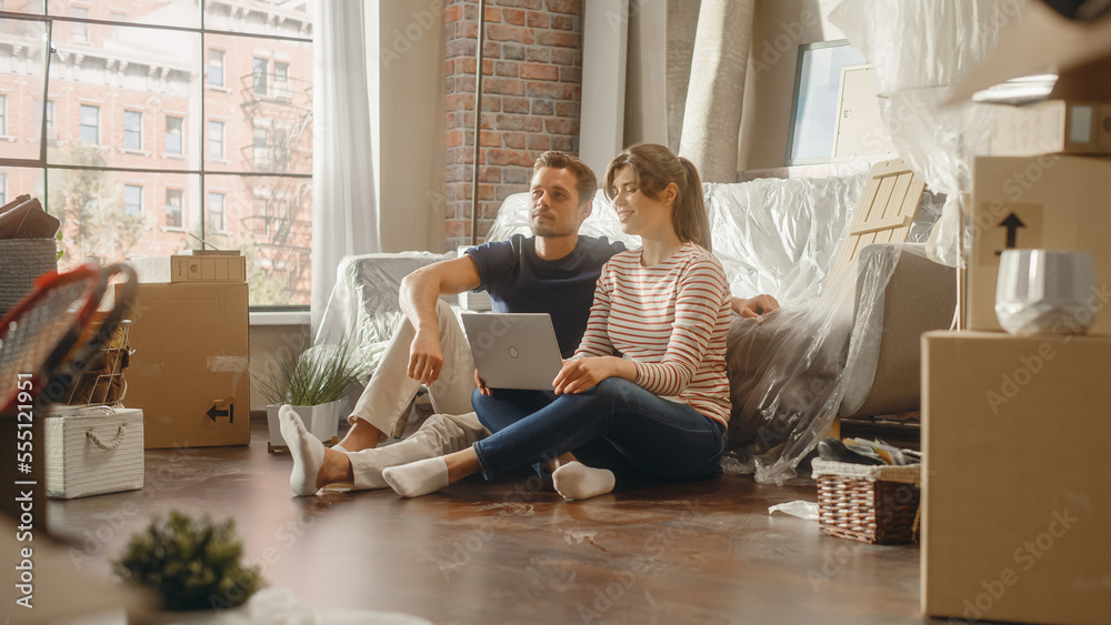 Moving in: Happy Young Homeowners: Couple Sitting on the Floor of their New Apartment Use Laptop Com