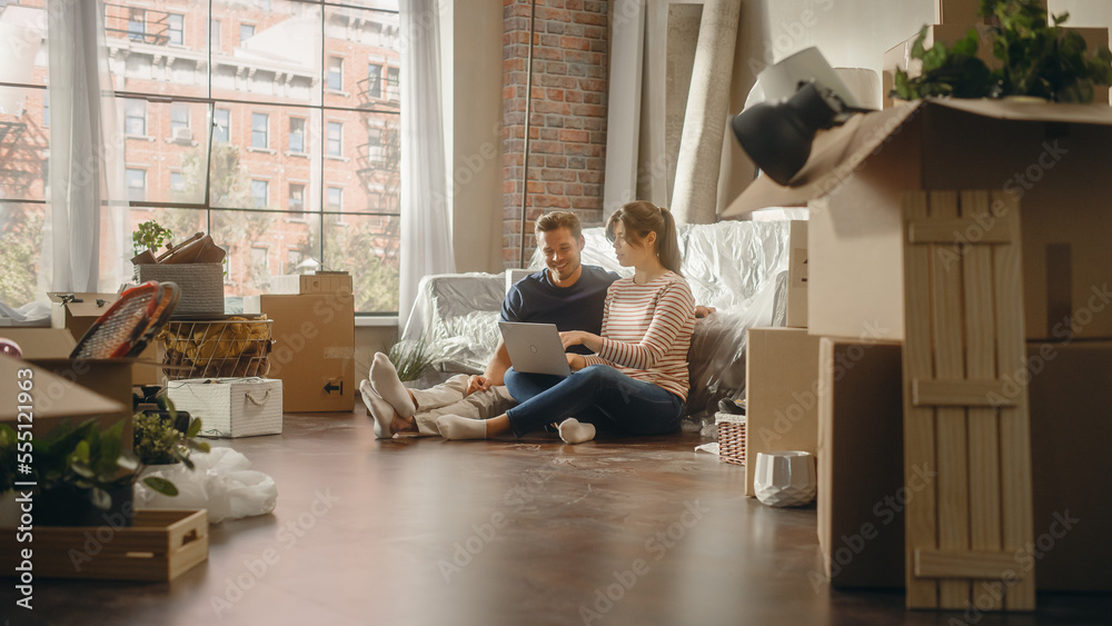 Moving Young Homeowners: Couple Sitting on the Floor of their New Apartment Use Laptop Computer for 