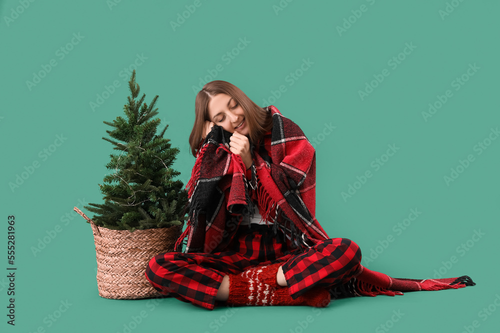 Young woman in pajamas with plaid and Christmas tree on green background