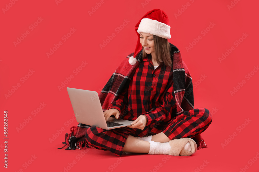 Young woman in Santa hat and pajamas using laptop on red background