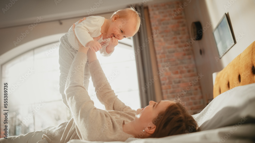 Loving Young Beautiful Mother Playing and Soothing Adorable Baby in Bed. Smiling Caring Mom and Cute