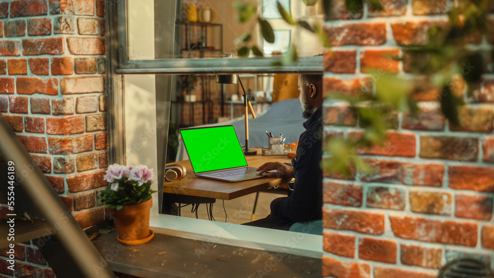 African Man Working on a Laptop Computer with Green Screen Mock Up Display while Sitting at a Table 