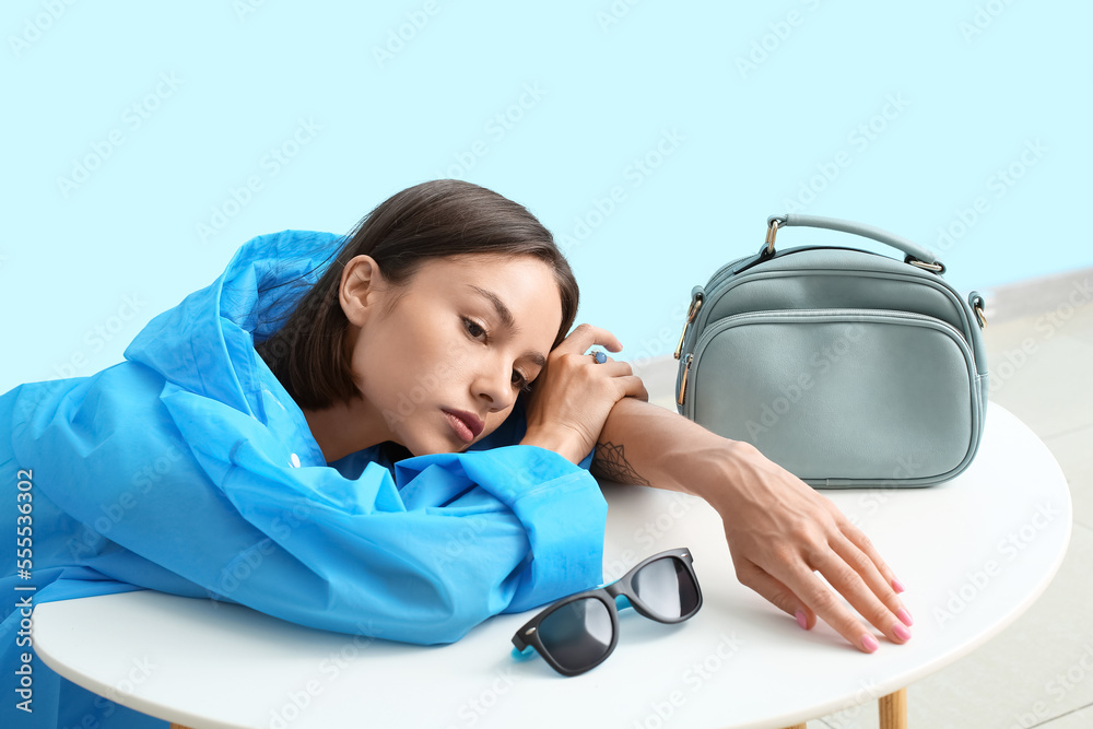 Young fashionable woman lying at table with stylish handbag and sunglasses against color background