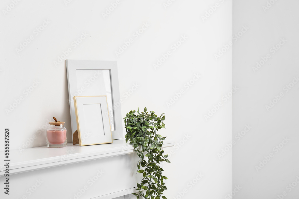 Blank photo frames, candle and houseplant on mantelpiece near white wall