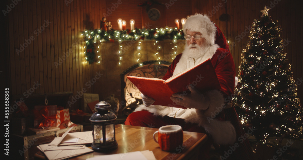 Joyful santa clause sitting in his rocker in decorated room, reading a book with red cover - holiday