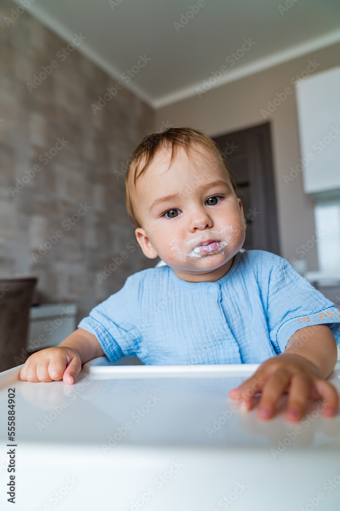 Baby eating food on kitchen. Grimy funny child eats sour cream in the chair.