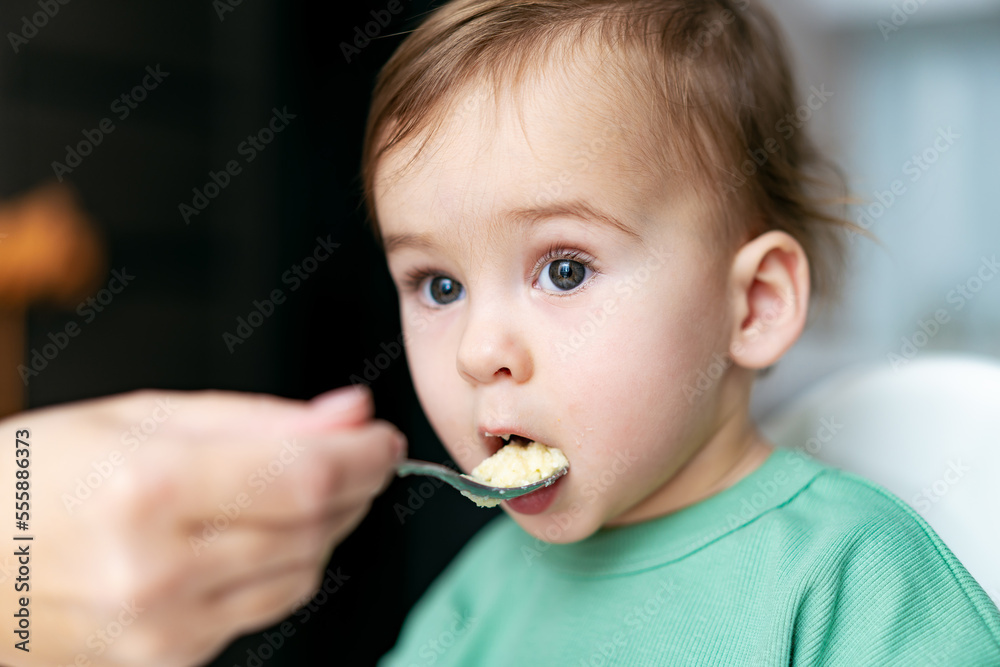 Mother feeding baby with spoon in kitchen. Funny baby one year old eating healthy food