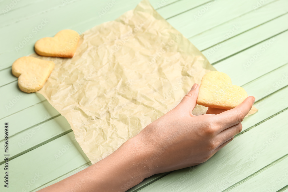 Female hand with heart shaped cookie and baking paper on green wooden background, closeup