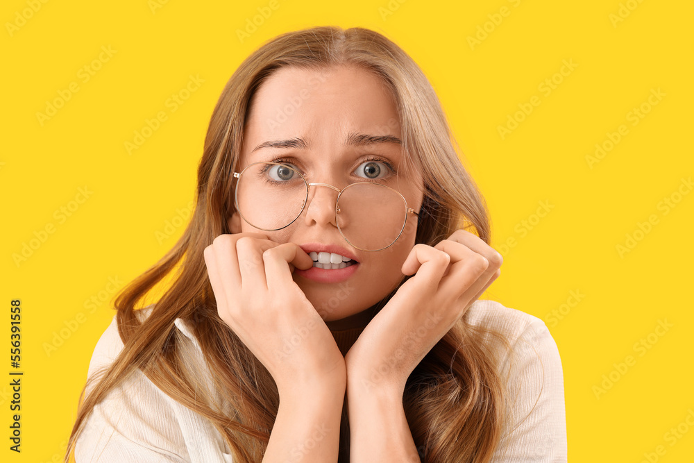 Young woman in eyeglasses biting nails on yellow background, closeup