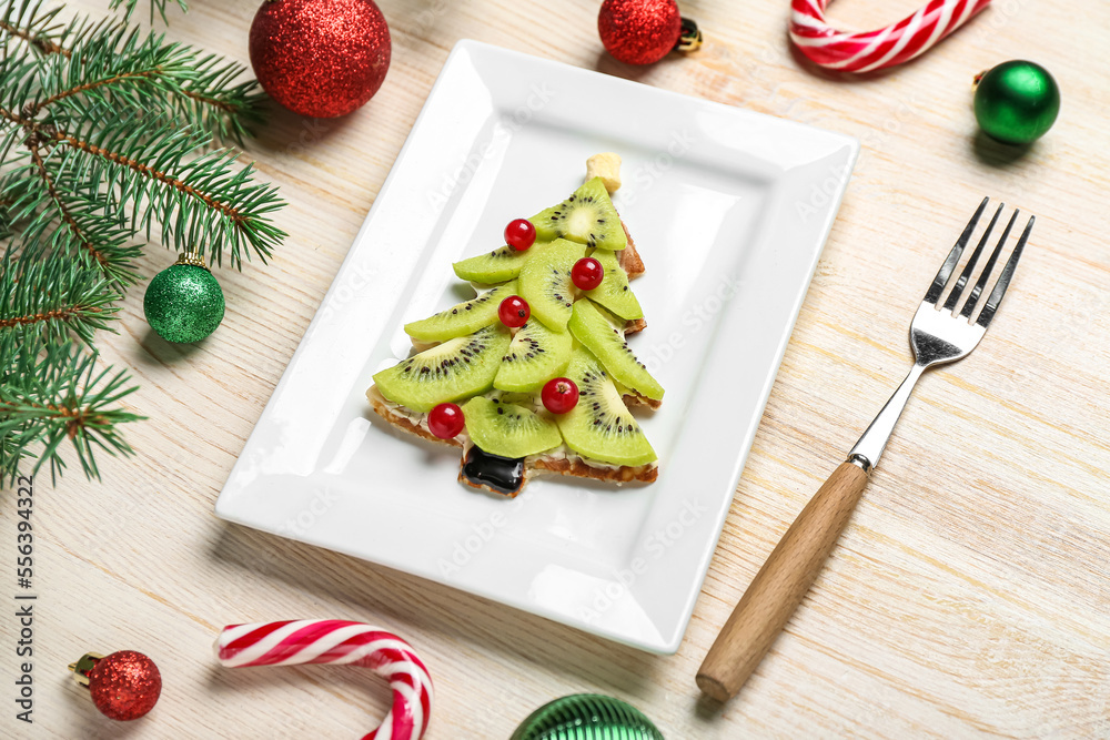 Plate of tasty pancake in shape of Christmas tree with fruits, fir branches and decorations on woode