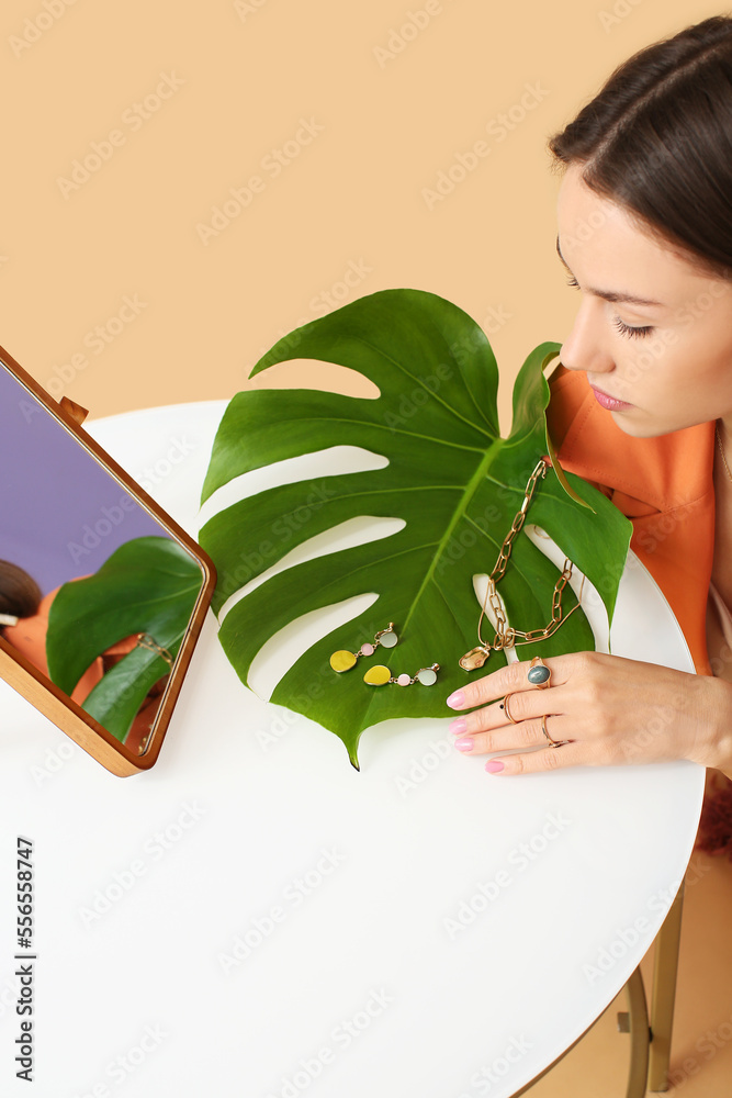 Young woman sitting at white table with stylish jewelry, palm leaf and mirror on color background