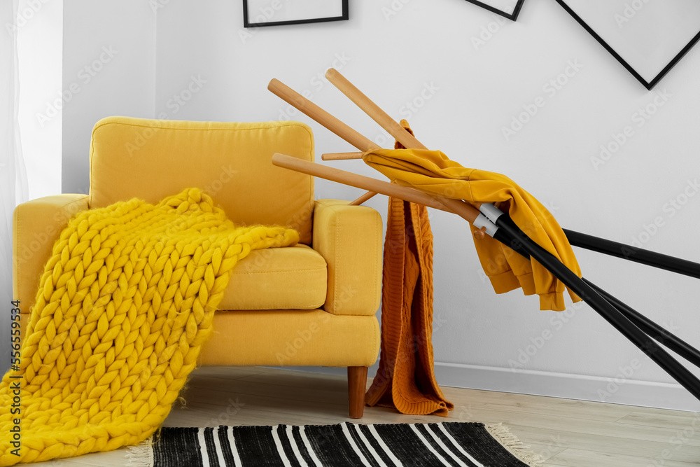 Yellow armchair with plaid and fallen clothes stand near light wall