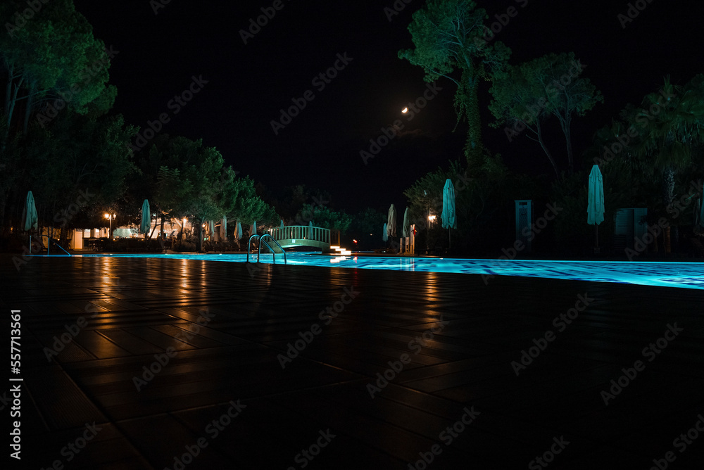 View of illuminated swimming pool surrounded with folded parasols and lush trees growing with sky in