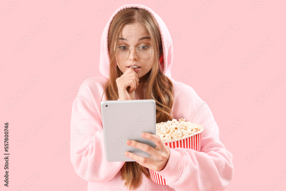 Young woman with tablet computer and popcorn biting nails on pink background