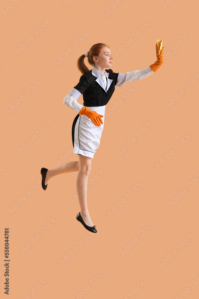 Young female chambermaid with rag jumping on beige background