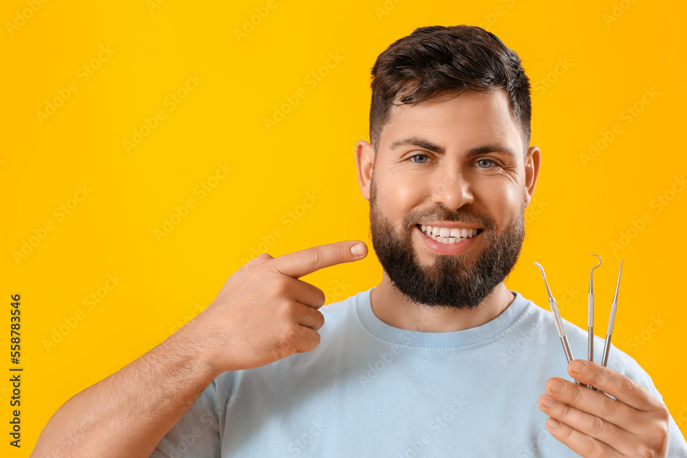 Young man with dental tools pointing at his teeth on yellow background, closeup