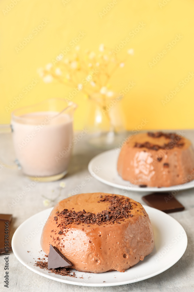 Plates with delicious pudding, chocolate and cup of milk on grey table near yellow wall