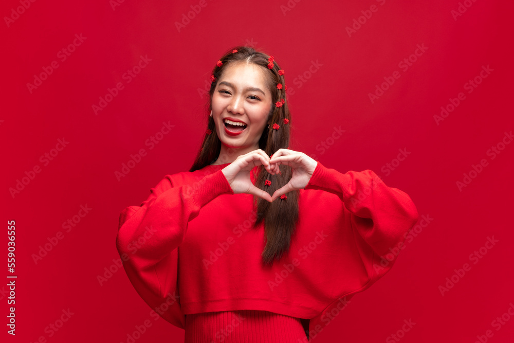 Young asian woman wearing red sweater dress shapes heart gesture on red background for Chinese new y