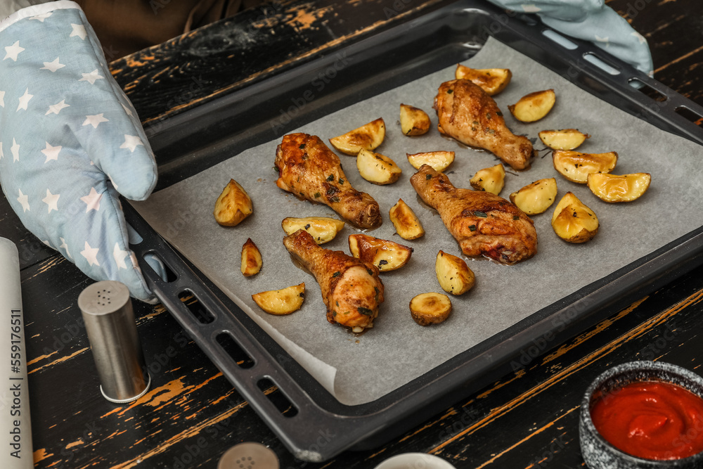 Woman holding baking tray with paper, tasty chicken legs and potato at dark wooden table, closeup