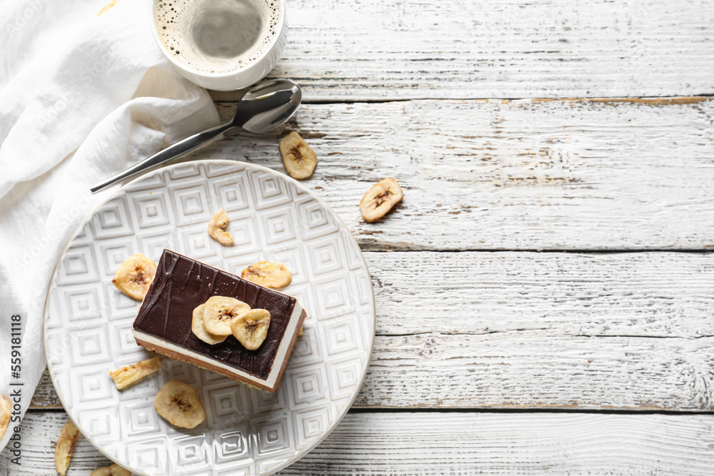 Plate with piece of birds milk cake, dried banana slices, cup of coffee and spoon on white wooden ba