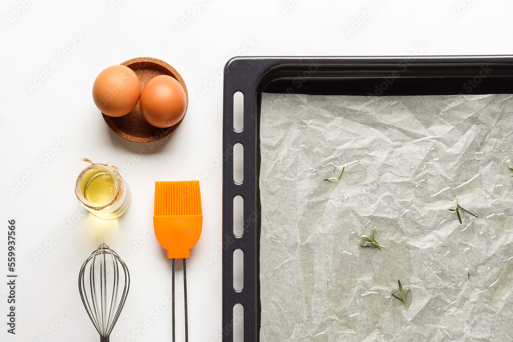 Baking tray with paper, eggs, oil, whisk and brush on white background